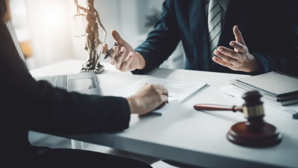 A legal discussion taking place with hands gesturing over a contract, with a small statue of Lady Justice on the table.