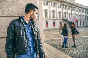 A man suspiciously watching two women from behind a wall in an urban setting.

