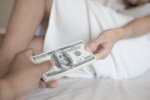Person handing cash to someone on a bed, partially covered by a blanket.