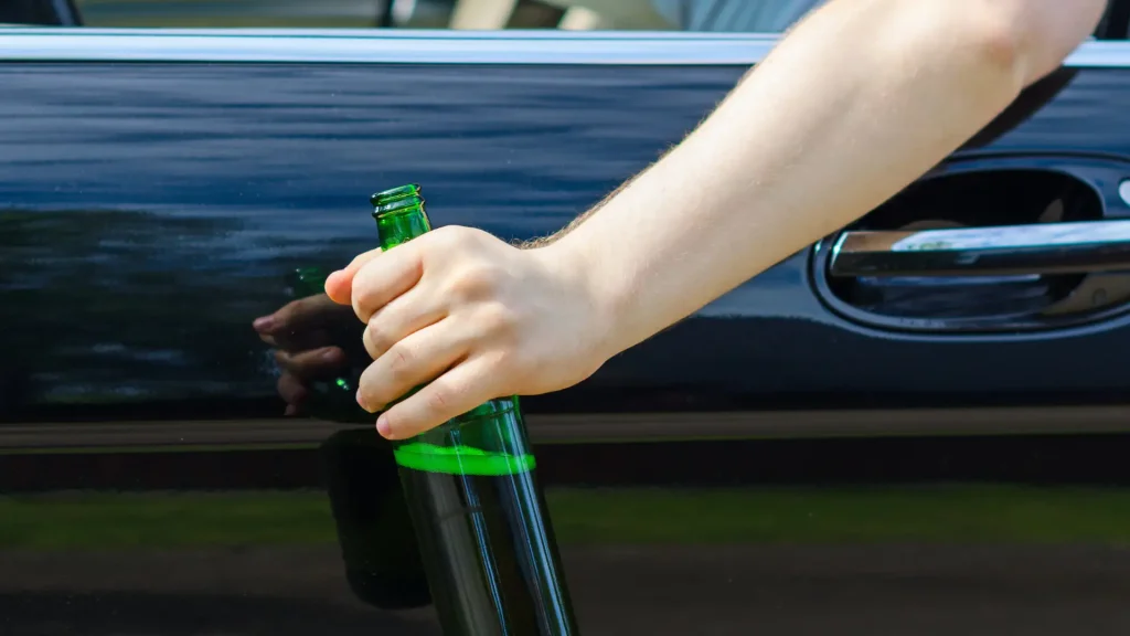 A person holding a green beer bottle near a car door, suggesting drinking and driving.